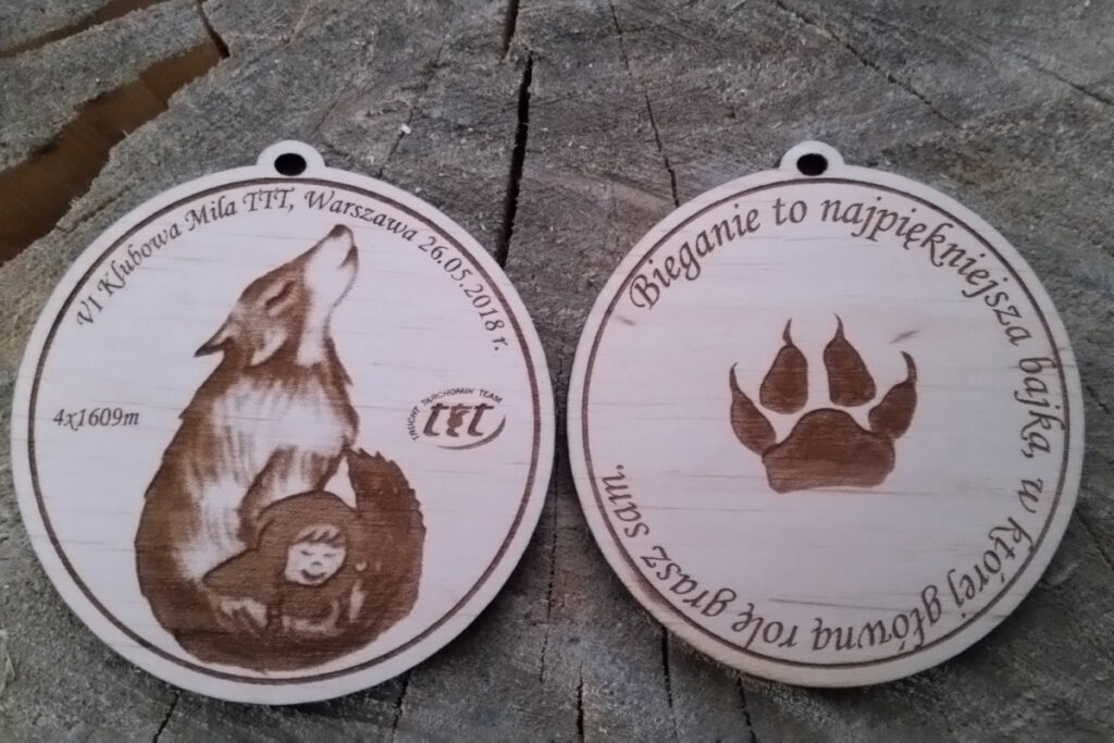 Dwustronny drewniany medal sportowy na wydarzenie Klubowa Mila TTT, z precyzyjnym grawerem laserowym, ażurowymi wycięciami na krawędziach i biało-czerwoną wstążką, ułożony na ciemnym, drewnianym blacie.