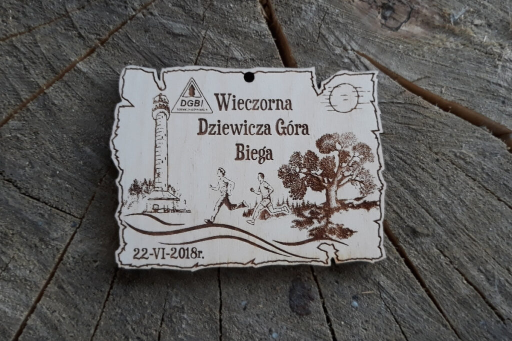 Nieregularnych kształtów drewniany medal z Biegu Wieczorna Dziewicza Góra Biega z nagrawerowanym motywem drzewa, biegaczy i wieży na Dziewiczej Górze.