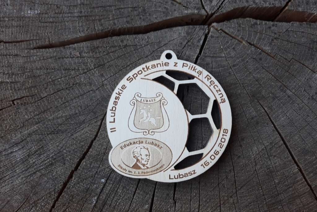 Drewniany medal ażurowy na II Lubaskie Spotkanie z Piłką Ręczną z wyciętyą ażurowo częścią piłki oraz logiem i napisami.