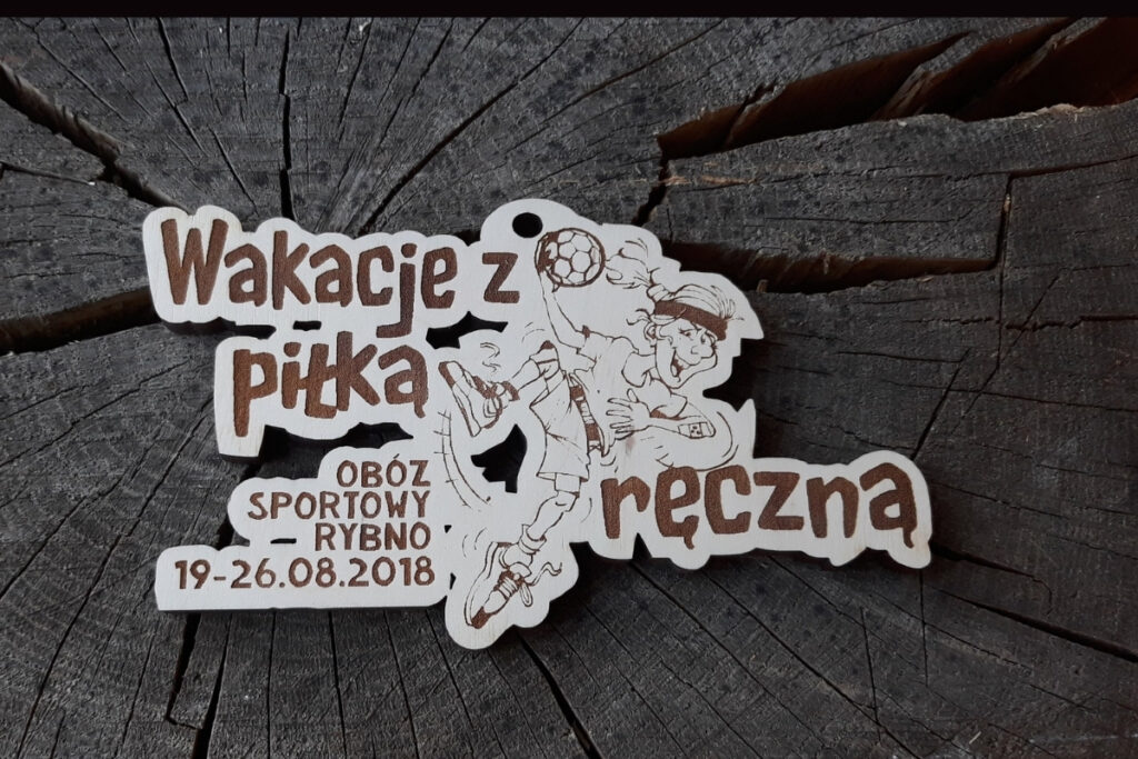 Drewniany medal pamiątkowy z obozu sportowego "Wakacje z piłką ręczną" w Rybnie z śmiesznym grawerem zawodnika i piłki ręcznej. Medal bardzo nieregularnych kształtów, poobcinany laserowo po ob rysach napisów i konturu zawodnika.