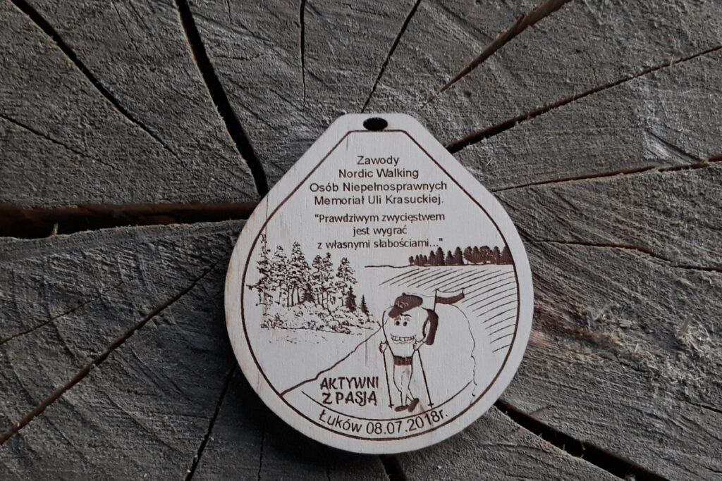 Drewniany medal pamiątkowy na Zawody Nordic Walking Osób Niepełnosprawnych z grawerem krajobrazu i logiem zawodów.
