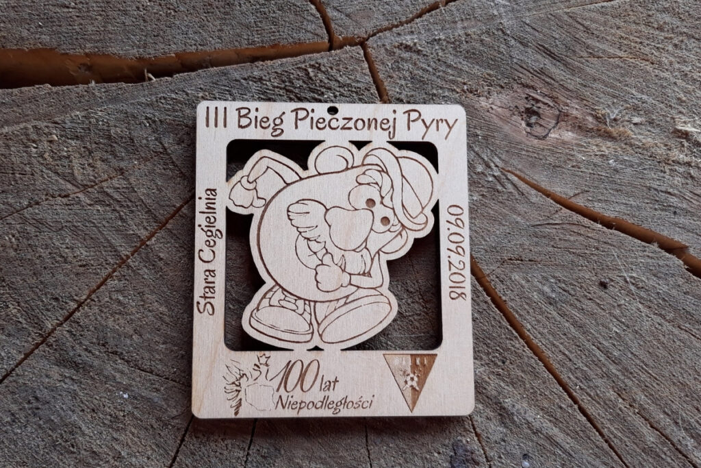 Kwadratowy drewniany medal na Bieg Pieczonej Pyry z ażurowym wycięciem biegnącego ziemniaka.