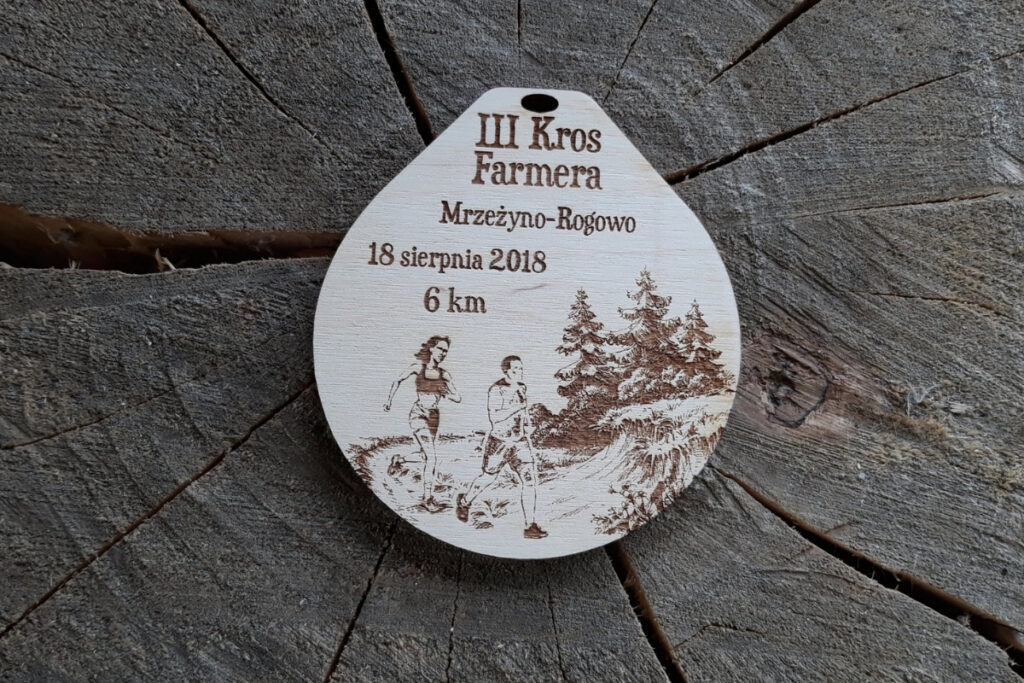 Drewniany medal na III Kros Farmera z grawerem biegaczy na ścieżce obok kępy z drzewami.