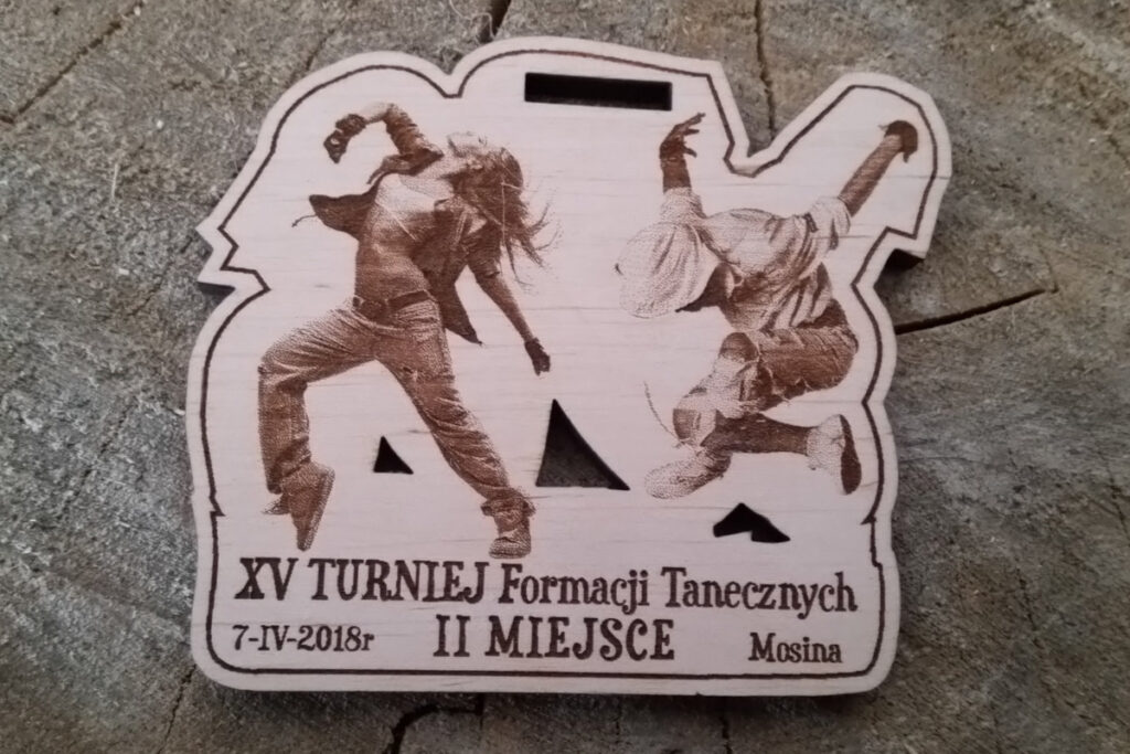 Nieregularnych kształtów drewniany medal z XV Turnieju Formacji Tanecznych w Mosinie, ozdobiony grawerem przedstawiającym sylwetkę tancerek w dynamicznej pozie.