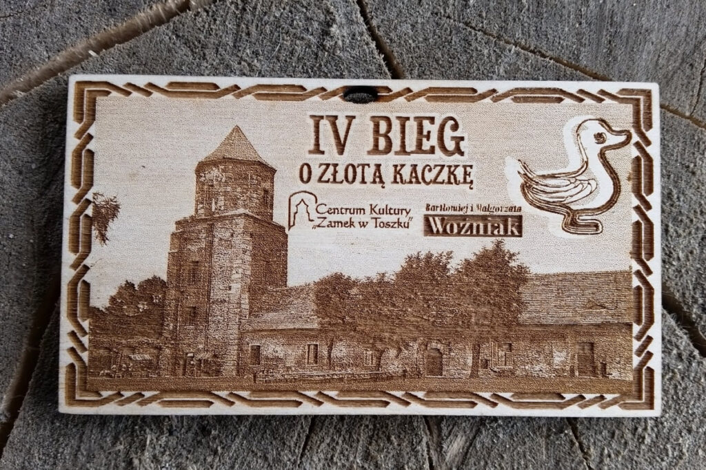 Prostokątny drewniany medal z grawerem Zamku w Toszku, postacią kaczki i napisem „IV Bieg o Złotą Kaczkę”.