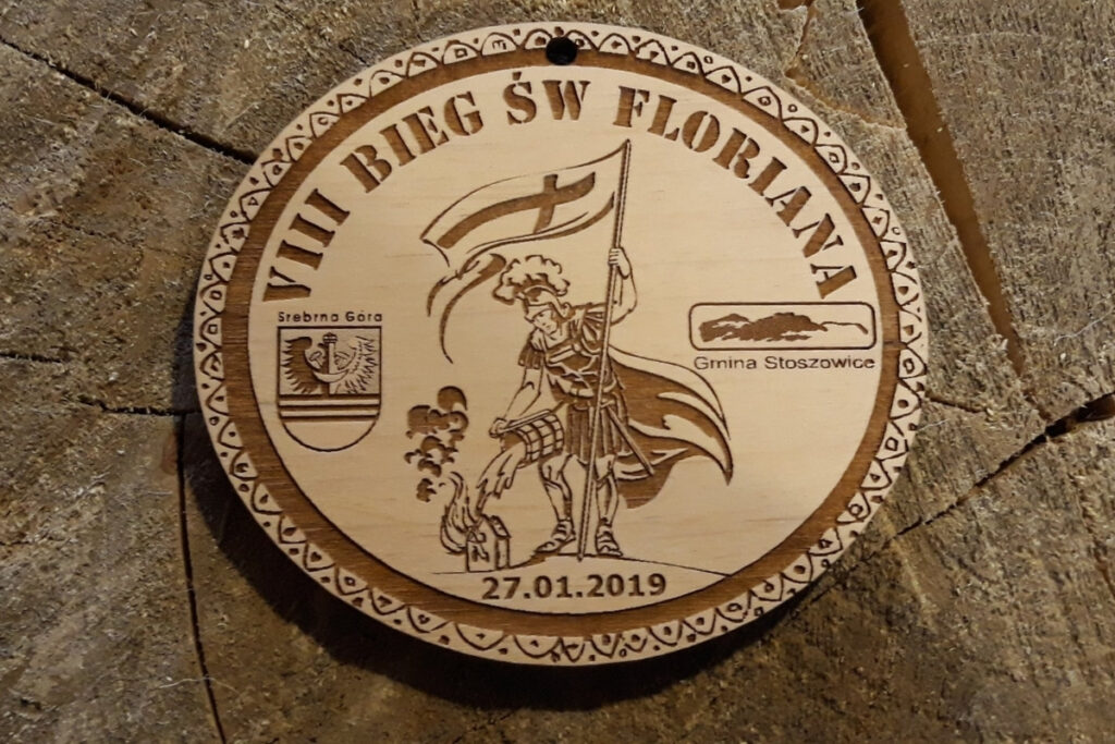 Drewniany medal na VIII Bieg św. Floriana z grawerem postaci Św Floriana i logotypami.