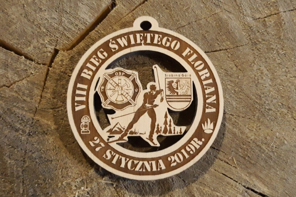 Okrągły, ażurowy medal wykonany z jasnego drewna na VIII Bieg Świętego Floriana z wyciętą sylwetką narciarza biegowego.