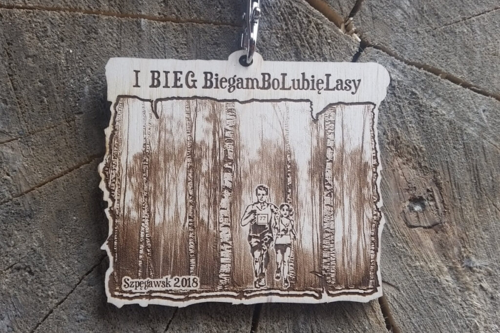 Drewniany medal o nieregularnym kształcie z grafiką biegaczy w lesie i napisem I Bieg BiegamBoLubięLasy w Szpęgawsku