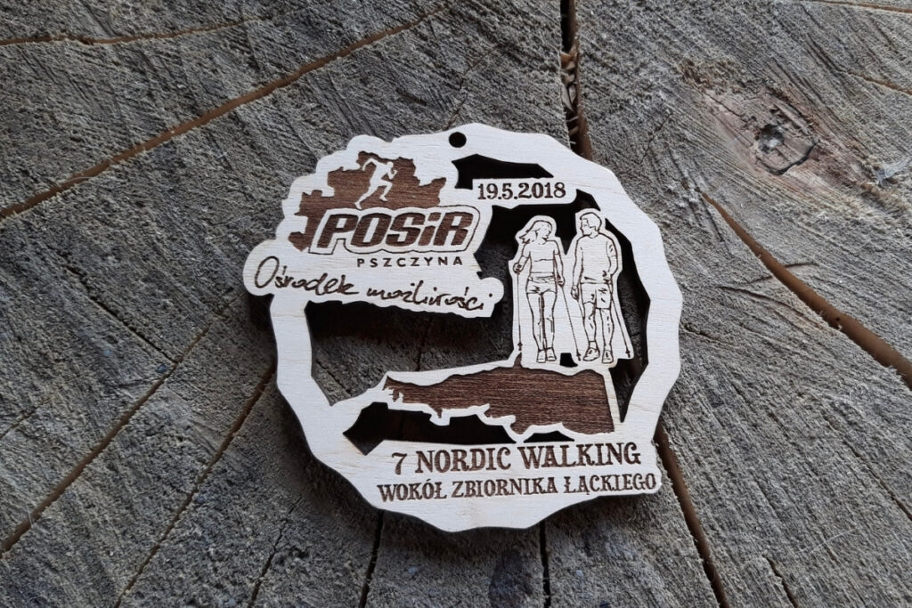 Drewniany, ażurowy medal pamiątkowy z 7. Nordic Walking wokół Zbiornika Łąckiego z wyciętymi ażurowo sylwetkami kobiety i mężczyzny maszerującymi z kijkami.