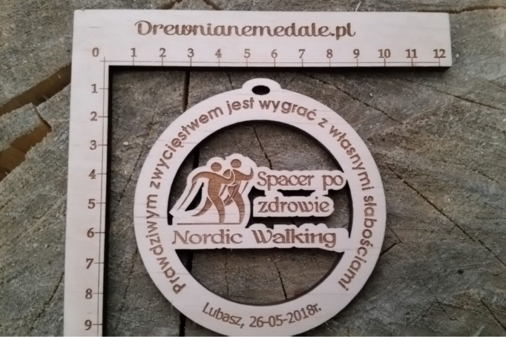 Ażurowy drewniany medal z grawerem „Spacer po zdrowie” przedstawiający postać z kijkami do nordic walking na tle lasu.
