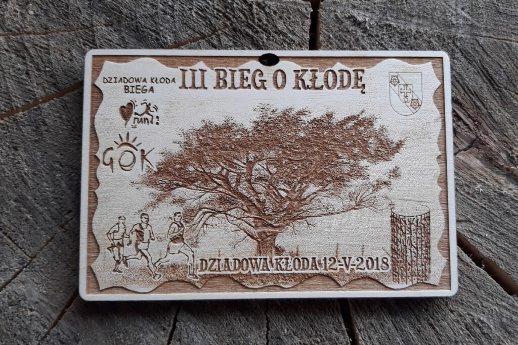 Prostokątny drewniany medal na III Bieg o Kłodę w Dziadowej Kłodzie z grawerowaną grafiką biegaczy, pnia i korony drzewa w ozdobnej ramce.