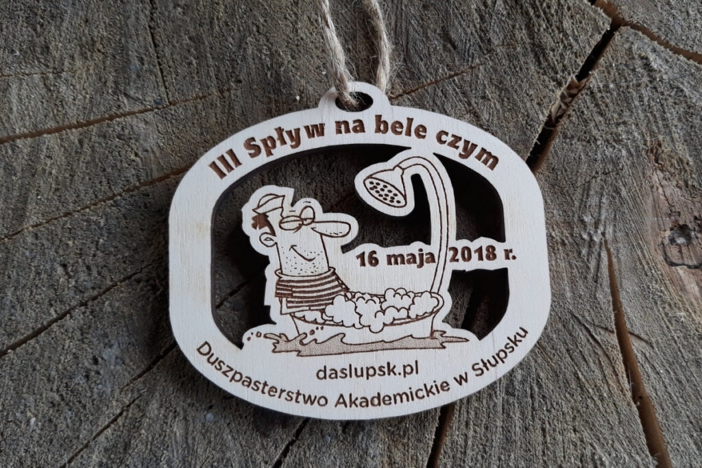 Drewniany, ażurowy medal na III Spływ na Bele Czym z wyciętym motywem śmiesznej postaci w bańce pod prysznicem.