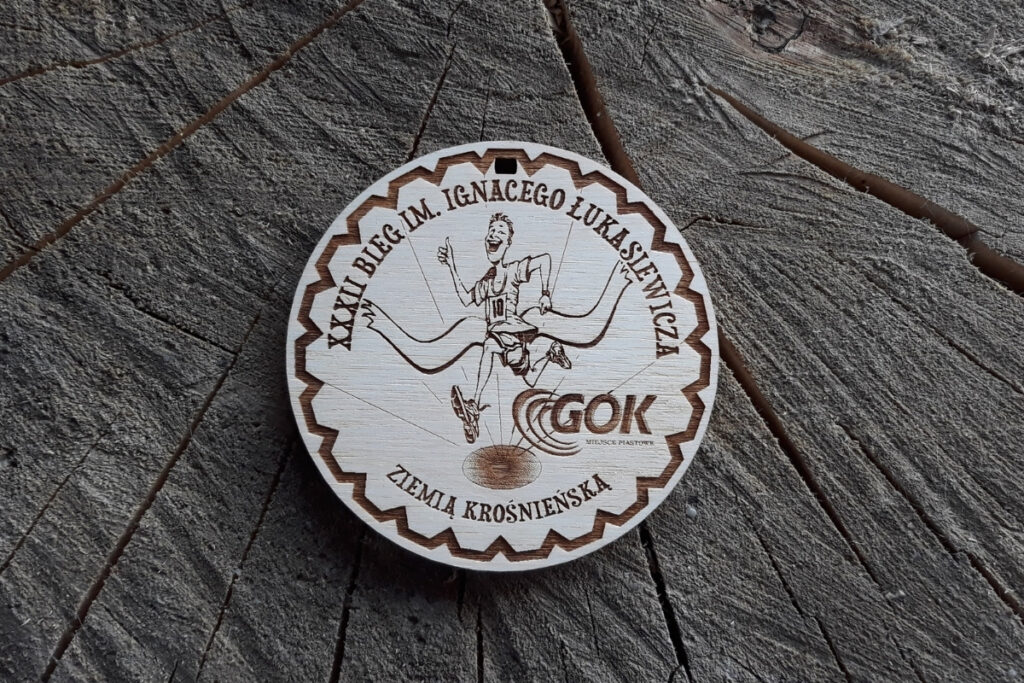 Drewniany medal pamiątkowy XXXII Biegu im. Ignacego Łukasiewicza z wygrawerowanym biegaczem ze wstęgą.