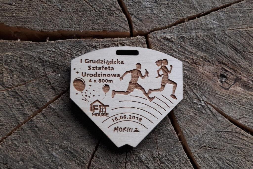 Zdjęcie drewnianego medalu puzzle z I Grudziądzkiej Sztafety Urodzinowej. Medal składa się z czterech dopasowanych do siebie elementów, które po złączeniu tworzą prostokąt z grawerowaną tabliczkę.