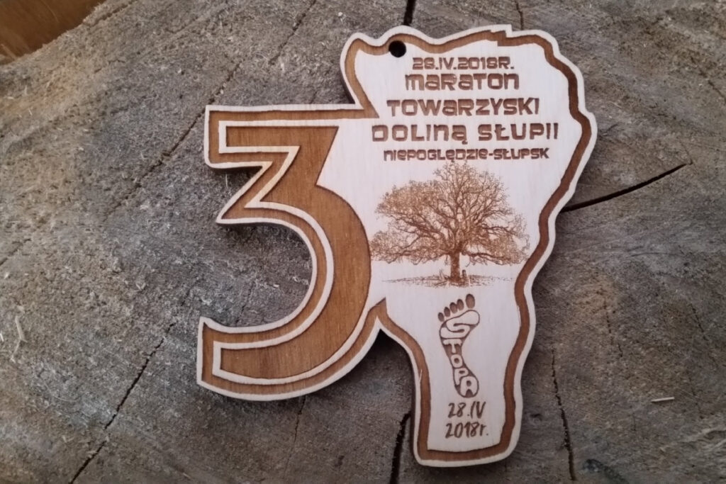 Drewniany medal w kształcie tarczy z grawerem trasy Maratonu Towarzyskiego Dolina Słupi, przedstawiający rzekę, las i sylwetkę biegacza.
