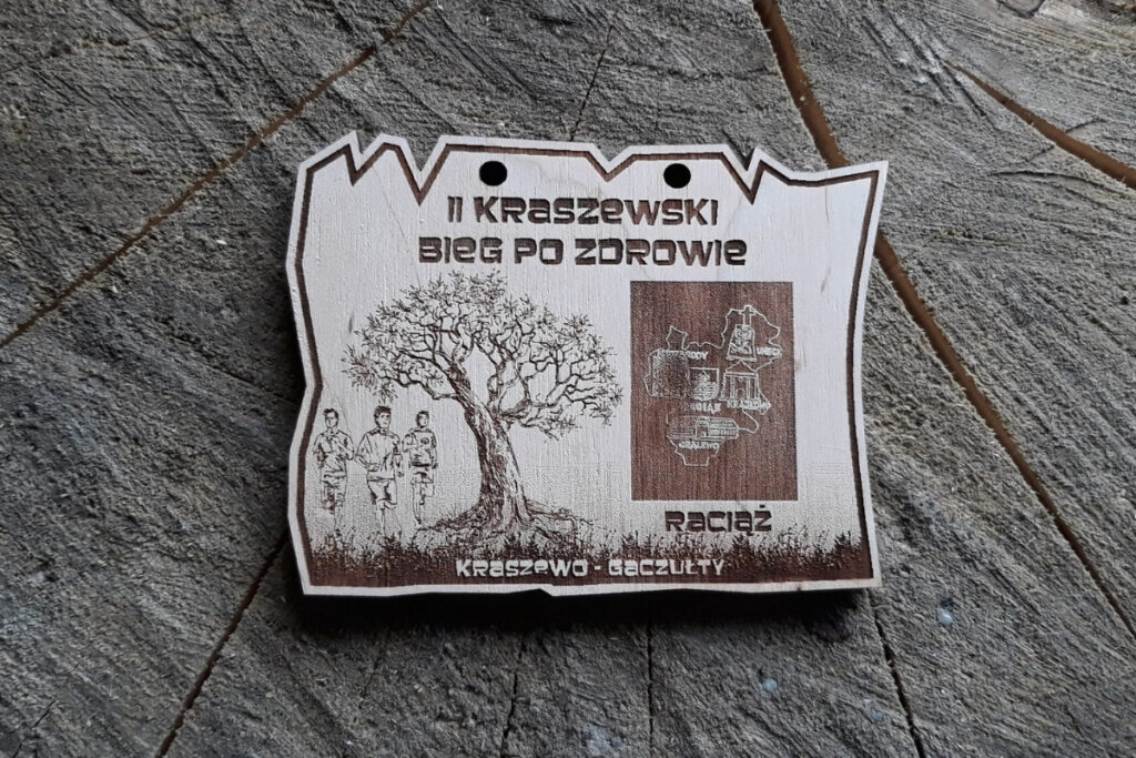 Ekologiczny prostokątny medal wykonany z jasnego drewna z nierównymi krawędziami. Na środku widnieje grawerowane sylwetki biegaczy oraz napis „II Kraszewski Bieg po Zdrowie”.