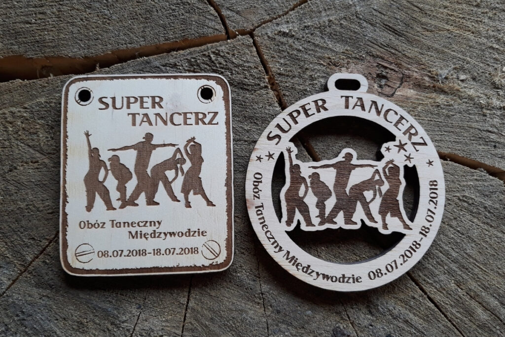 Drewniane medale z grawerem przedstawiającym sylwetkę tancerki w dynamicznej pozie. Na medalu widnieje napis 'Super Tancerz' oraz informacja o obozie tanecznym w Międzywodziu.