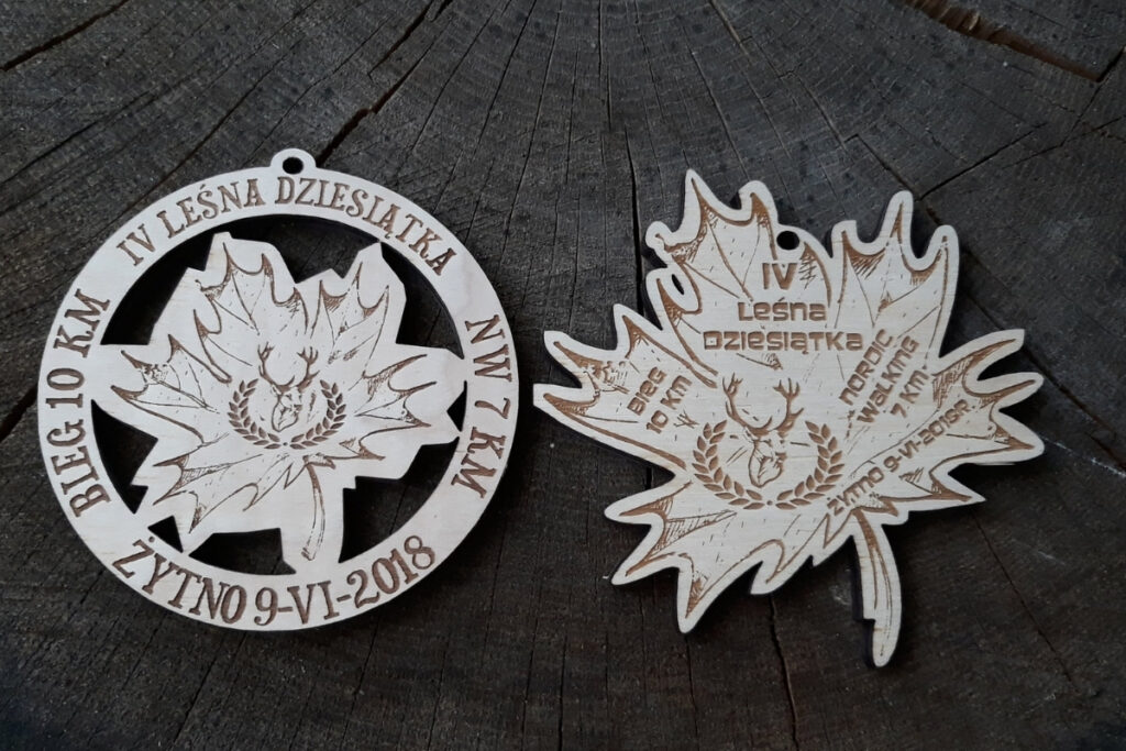 Drewniany medal pamiątkowy z biegu IV Leśna Dziesiątka Żytno 2018 z grawerem liścia. Na zdjęciu są 2 medale, jeden okrągły z ażurowo wyciętym liściem, drugi - sam liść o nieregularnych kształtach.