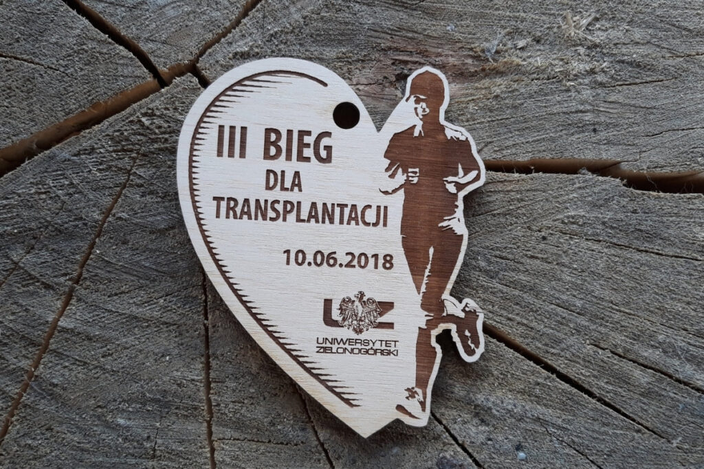 Drewniany medal pamiątkowy na III Bieg dla Transplantacji 2018 w kształcie pół-serca, w które wbiega wygrawerowany biegacz.