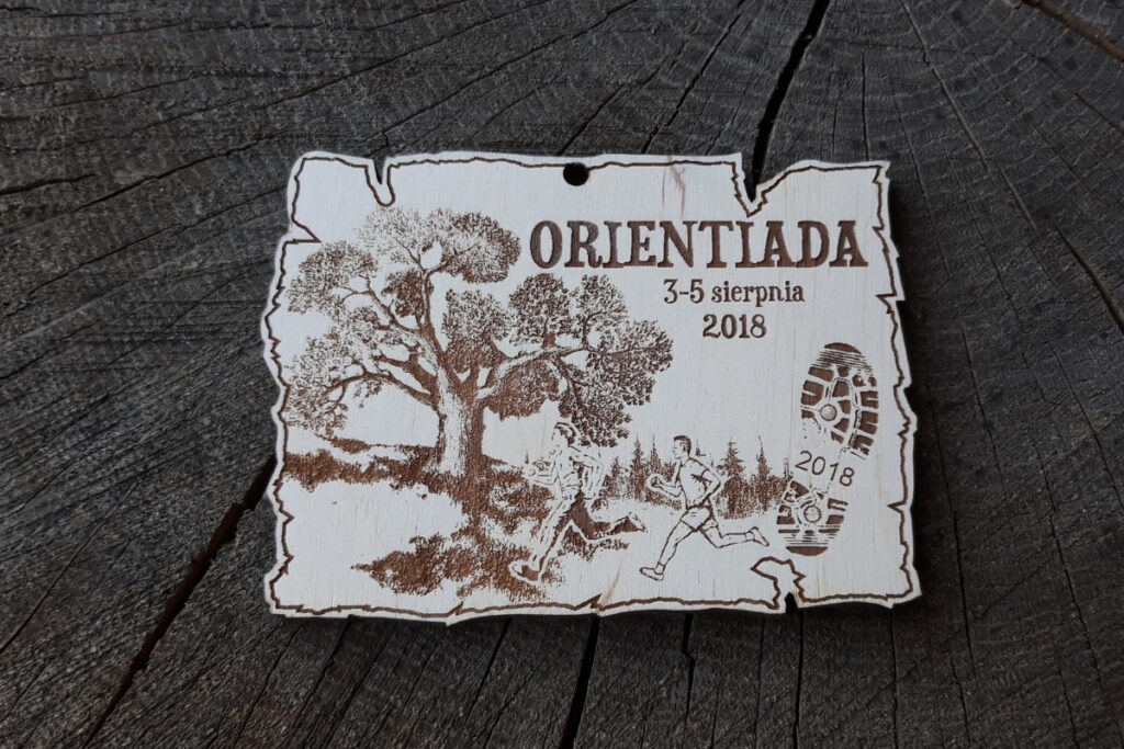 Drewniany prostokątny medal Orientiada 2018 na bieg na orientację z grawerem biegnących postaci, drzewem, butem oraz dużym napisem ORIENTIADA.