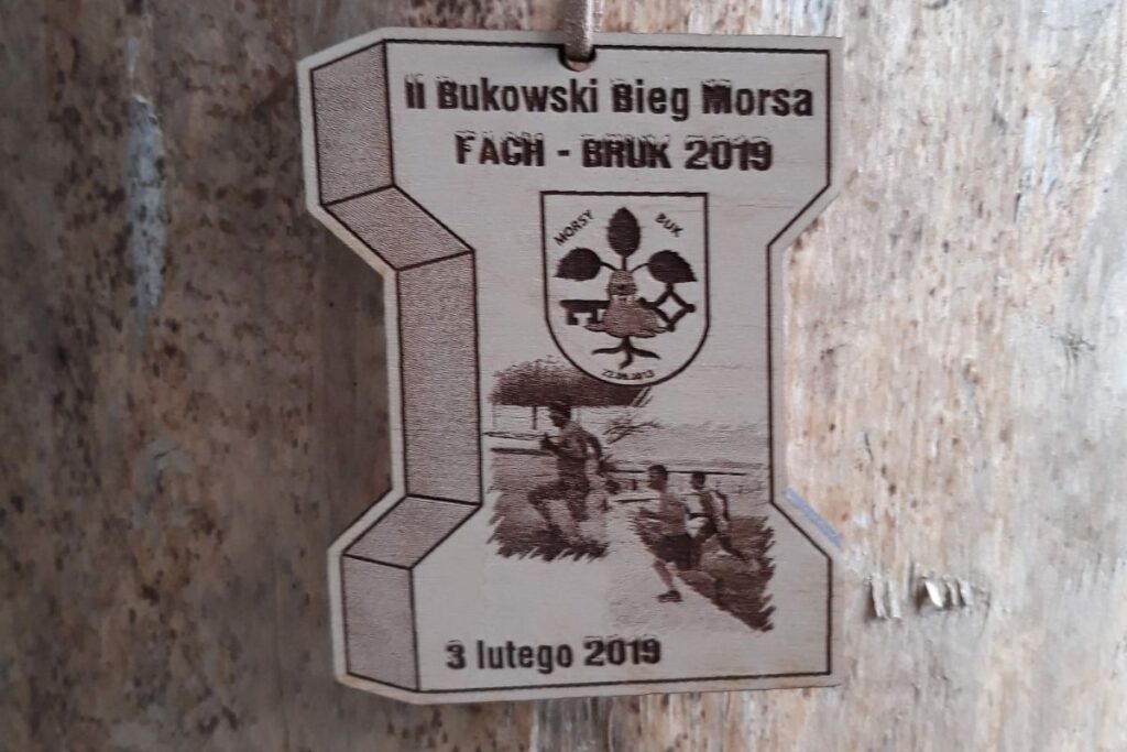 Drewniany medal w kształcie sześciennej kostki brukowej z grawerem II Bukowski Bieg Morsa i grafiką zdjęć osób morsujących.