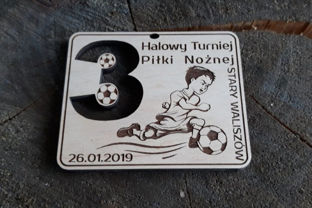 Drewniany medal sportowy na 3. Halowy Turniej Piłki Nożnej z grawerem młodego piłkarza w ruchu, piłką oraz ażurowo wyciętą cyfrą 3.