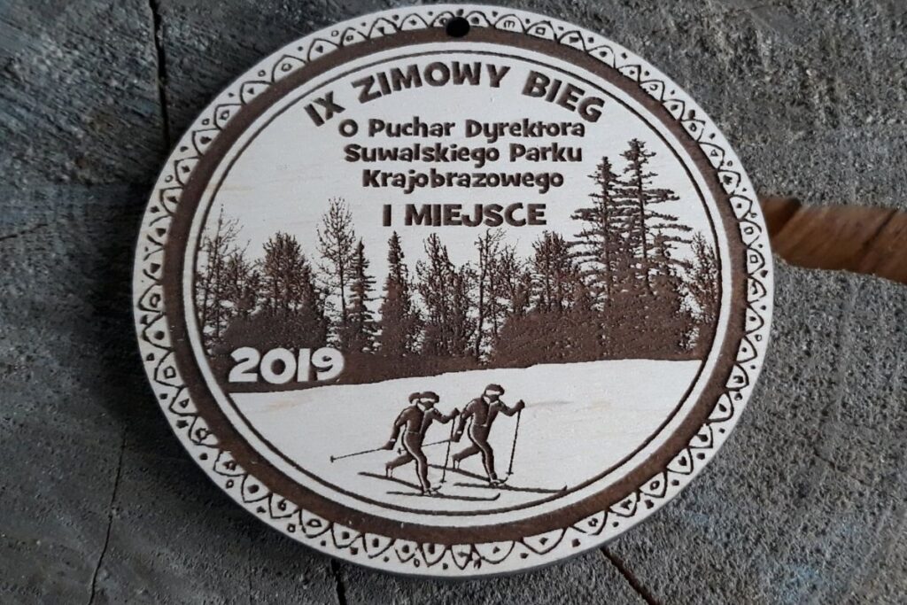 Drewniany medal pamiątkowy na zimowy bieg narciarski z narciarzy w biegu, w tle drzewa.