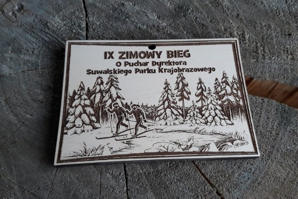 Drewniany medal pamiątkowy IV Zimowego Biegu o Puchar Dyrektora SPN z grawerem przedstawiającym zimowy krajobraz i logo Suwalskiego Parku Narodowego.