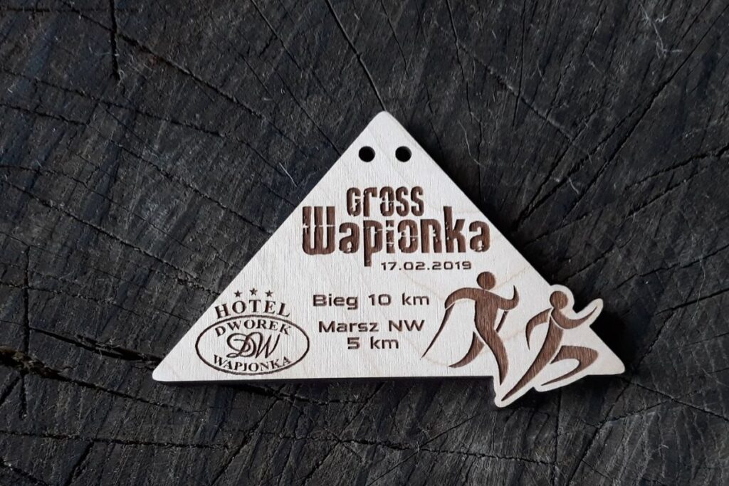 Drewniany medal na Cross Wapionka 2019 z grawerowanym motywem chodziarza Nordic Walking i biegacza.