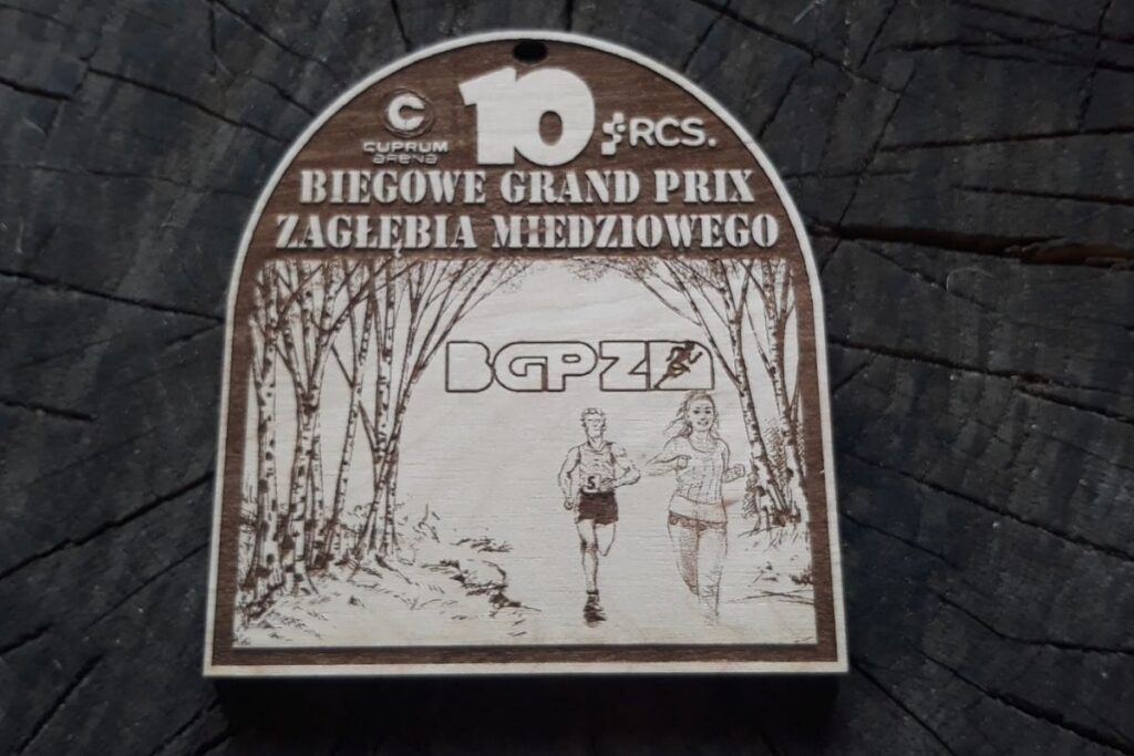 Propozycja drewnianego medalu na 10. Biegowe Grand prix Zagłębia Miedziowego. Drewniany medal z cyfrą 10 napisami i logiem.