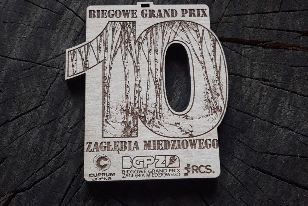 Propozycja drewnianego medalu na 10. Biegowe Grand prix Zagłębia Miedziowego. Drewniany medal z cyfrą 10 napisami i logiem.