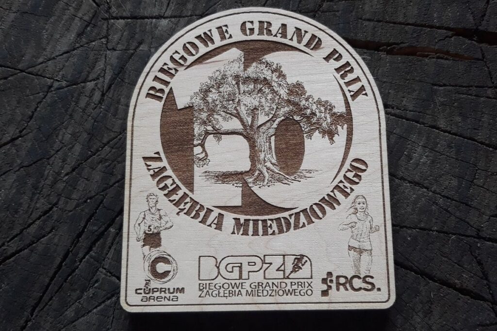 Propozycja drewnianego medalu na 10. Biegowe Grand prix Zagłębia Miedziowego. Drewniany medal z cyfrą 10 napisami i logiem.