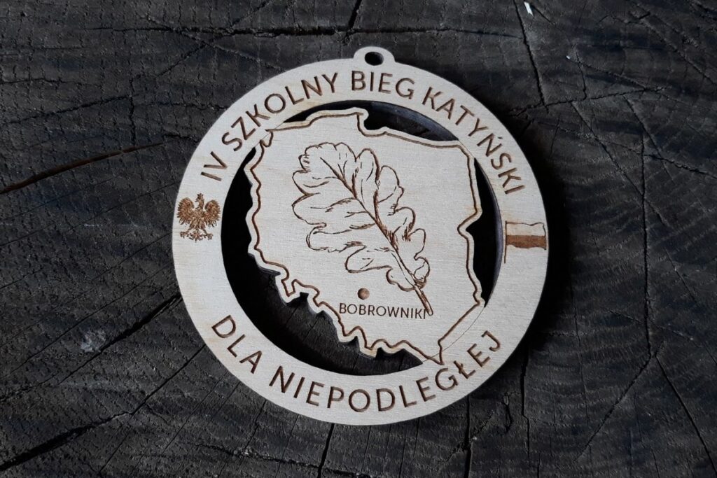 Okrągły medal z drewna z ażurowymi wycięciami, grawerem liścia w konturze Polski oraz zaznaczoną miejscowością Bobrowniki.