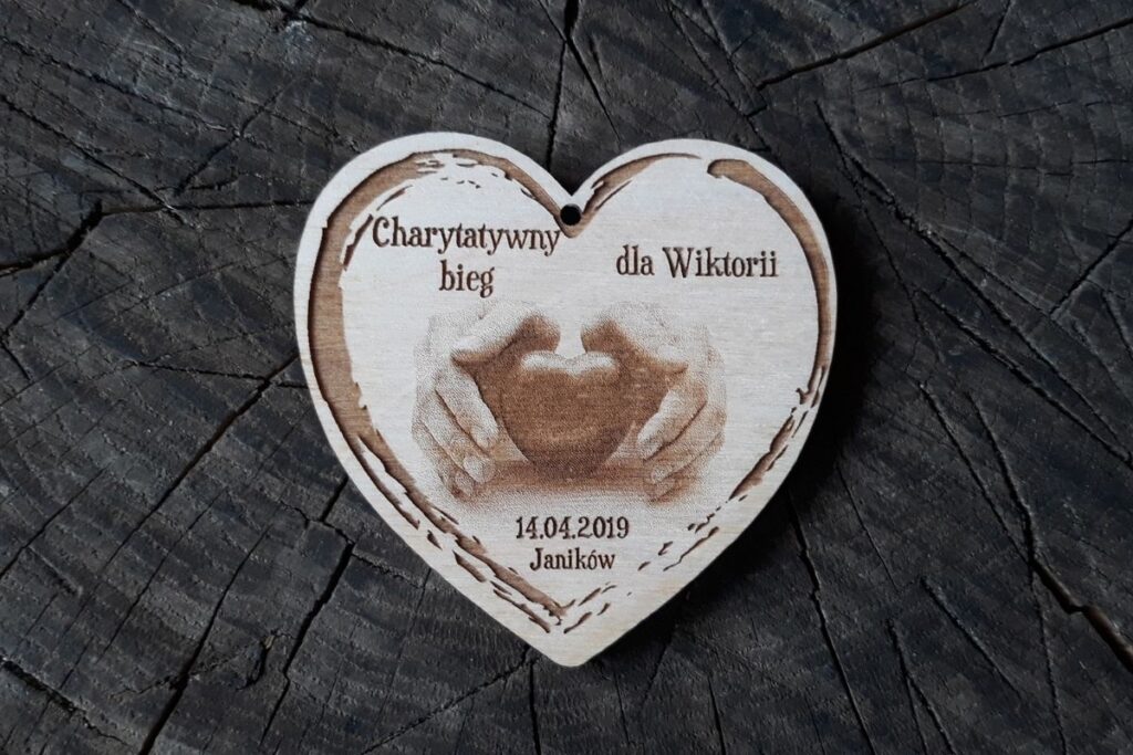 Drewniany medal w kształcie serca z wygrawerowanymi dłońmi obejmującymi serce i napisem charytatywnym.