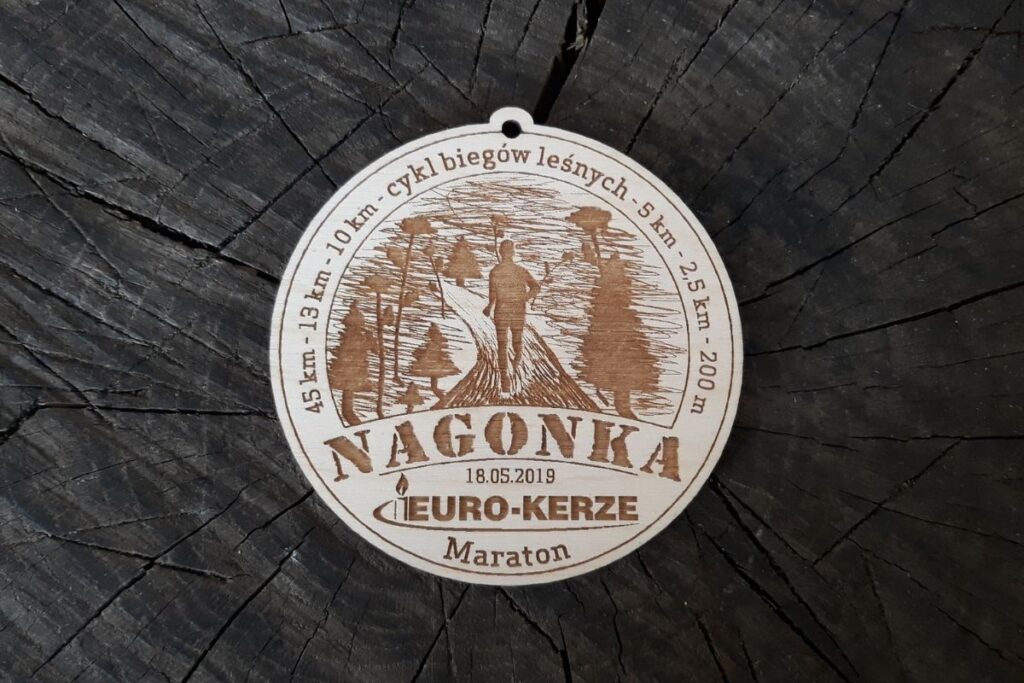 Okrągły drewniany medal z biegu Nagonka przedstawiający grawerowaną postać biegacza na krętej leśnej ścieżce wśród drzew.