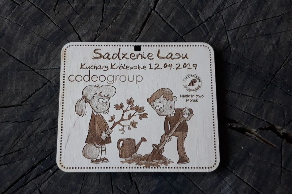Ekologiczny medal z naturalnego drewna upamiętniający akcję sadzenia drzew, z grawerem osób sadzących drzewka i logotypem.