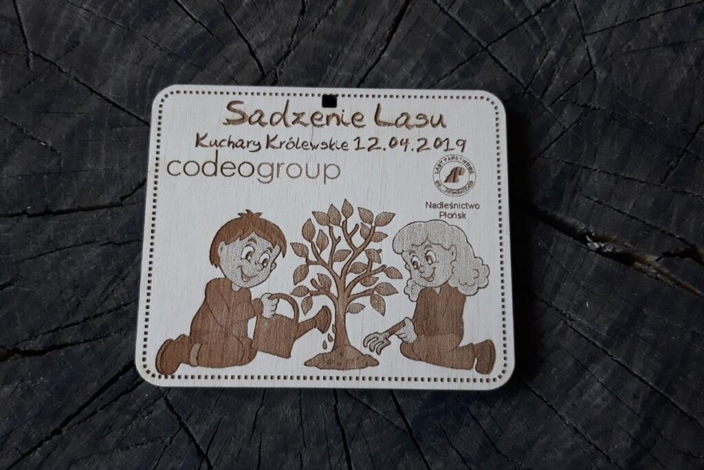 Ekologiczny medal z naturalnego drewna upamiętniający akcję sadzenia drzew, z grawerem osób sadzących drzewka i logotypem.