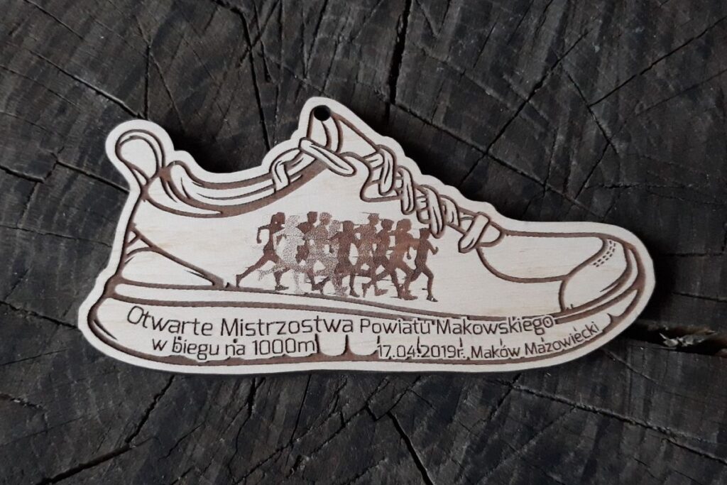 Drewniany medal w kształcie buta sportowego z wygrawerowanym napisem Mistrzostwa Powiatu Makowskiego oraz herbem powiatu.