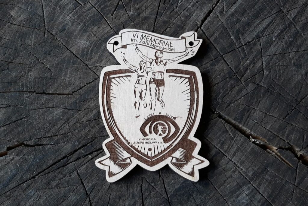 Drewniany medal w kształcie tarczy z wygrawerowanymi sylwetkami biegaczy, napisem VI Memoriał im. Zofii Morawskiej i logo wydarzenia.