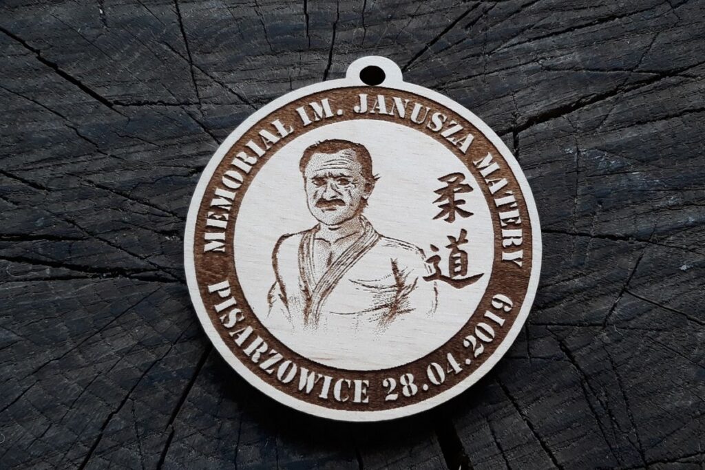 Okrągły drewniany medal pamiątkowy z grawerowanym portretem Janusza Matery, napisem Memoriał i datą wydarzenia.