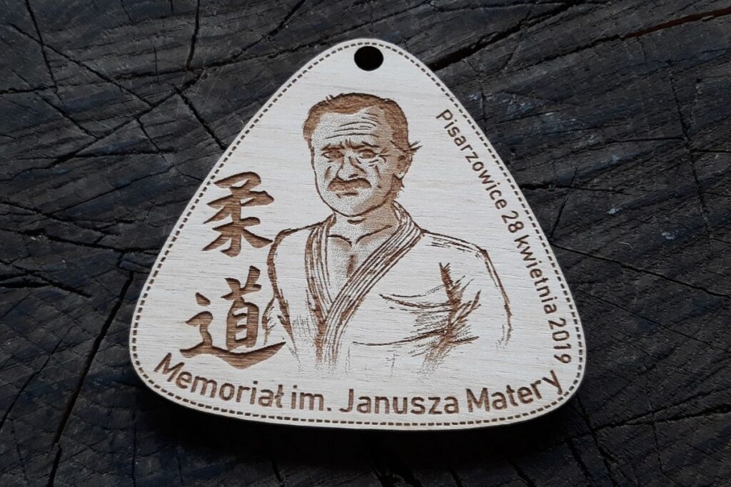Trójkątny drewniany medal pamiątkowy z grawerowanym portretem Janusza Matery, napisem Memoriał i datą wydarzenia.