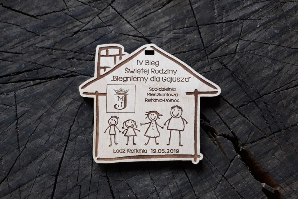 Drewniany medal w kształcie domku z wygrawerowaną uśmiechniętą rodziną trzymającą się za ręce, napisem IV Bieg Świętej Rodziny.