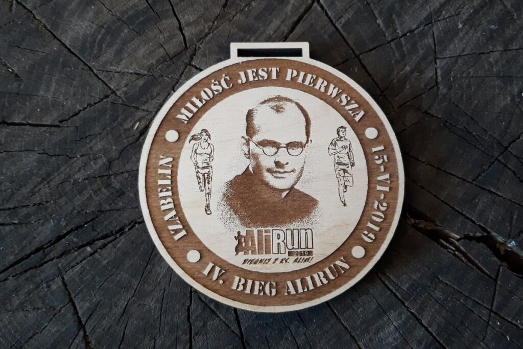 Okrągły drewniany medal z grawerowanym portretem ks. Aleksandra Fedorowicza, cytatem 'Miłość jest pierwsza' oraz datą i nazwą wydarzenia.