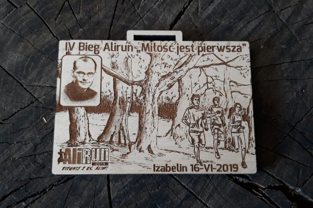 Prostokątny drewniany medal z grawerowanym wizerunkiem ks. Aleksandra Fedorowicza, napisem 'Miłość jest pierwsza' oraz datą wydarzenia.