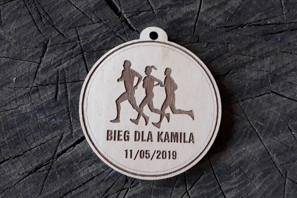 Okrągły drewniany medal z grawerowanymi sylwetkami biegnących osób i dużym napisem BIEG DLA KAMILKA.