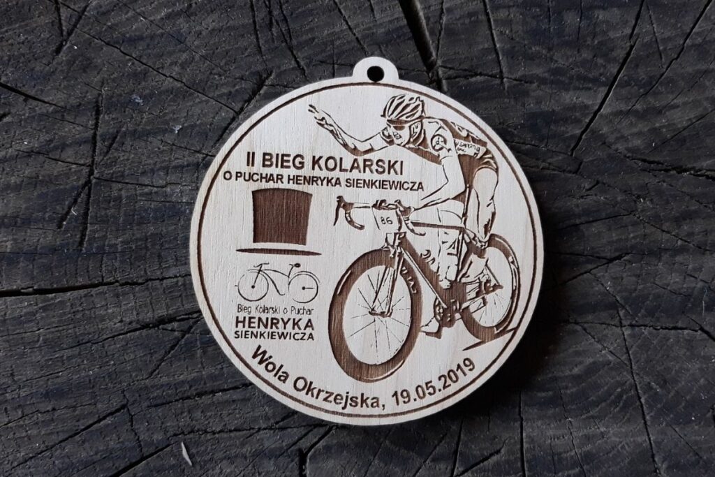 Okrągły drewniany medal z wygrawerowaną sylwetką kolarza w kasku na rowerze szosowym oraz napisem Bieg Kolarski.