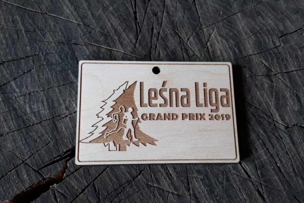 Drewniany medal z napisem Leśna Liga i osobą maszerującą oraz biegnącą na tle świerków.