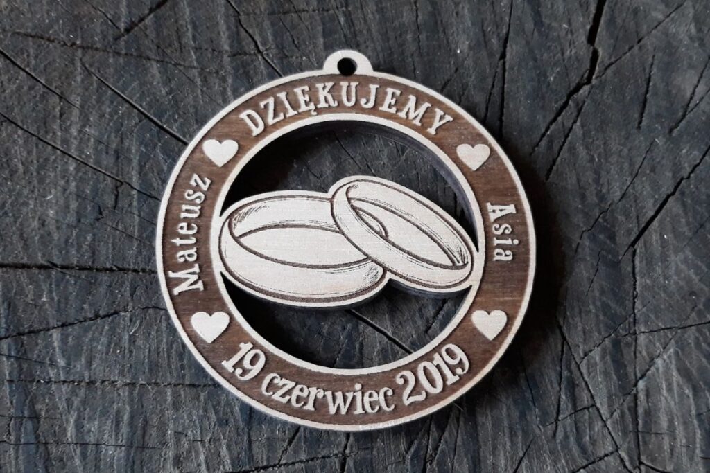 Okrągły drewniany medal weselny z wygrawerowanymi imionami Mateusz i Asia, napisem 'Dziękujemy za przybycie' oraz motywem ażurowo wyciętej obrączki.