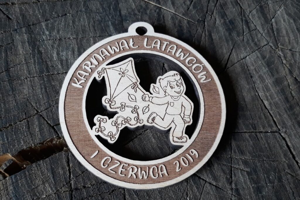 Drewniany medal ażurowy na Karnawał Latawców z wyciętym laserowo kształtem uśmiechniętego chłopczyka puszczającego latawiec.