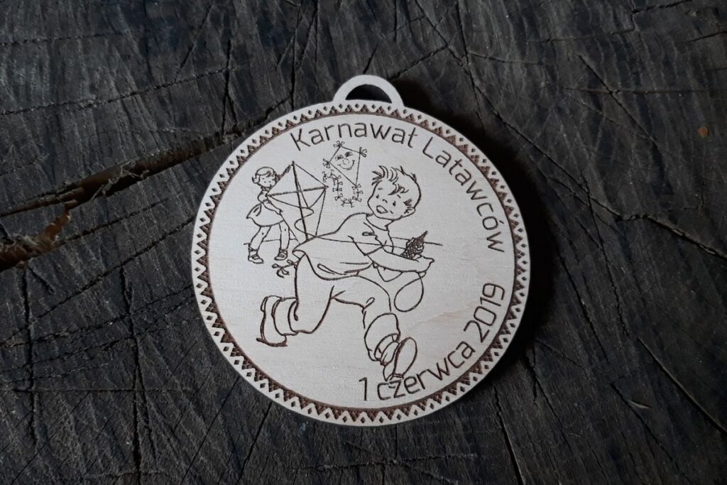 Okrągły drewniany medal na Karnawał Latawców z grawerem uśmiechniętego słońca wypuszczającego latawca z kokardkami.